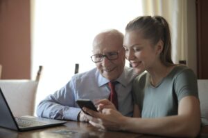 How To Keep Aging Parents At Home - Daughter sitting with senior father and watching a video on a smartphone