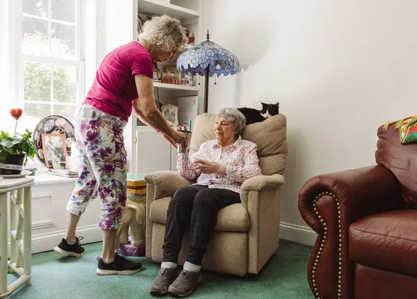 Aging In Place - daughter bringing drink to elderly mother sitting in a chair inside a house